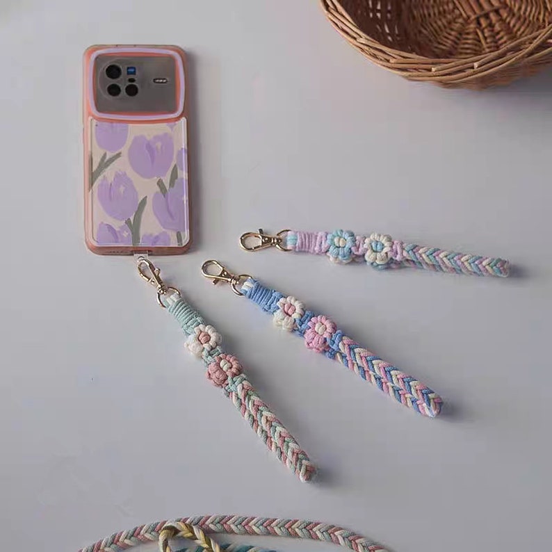 Pu&ograve; includere: Tre cinturini per telefono intrecciati colorati con ciondoli a forma di fiore. Ogni cinturino &egrave; realizzato con una diversa combinazione di colori di filo rosa, blu e bianco. I cinturini sono attaccati ad anelli di metallo dorati.