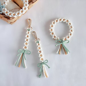 Könnte beinhalten: Drei handgefertigte Schlüsselanhänger in Form von Gänseblümchen mit grünen und gelben Quasten. Die Schlüsselanhänger bestehen aus weißem Garn und sind zu einer Gänseblümchenform zusammengefügt. Die Schlüsselanhänger hängen an einer weißen Oberfläche.