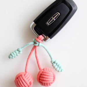 Op de afbeelding: Een zwarte autosleutel met een roze en lichtblauwe touwtjes sleutelhanger met twee roze geknoopte ballen.