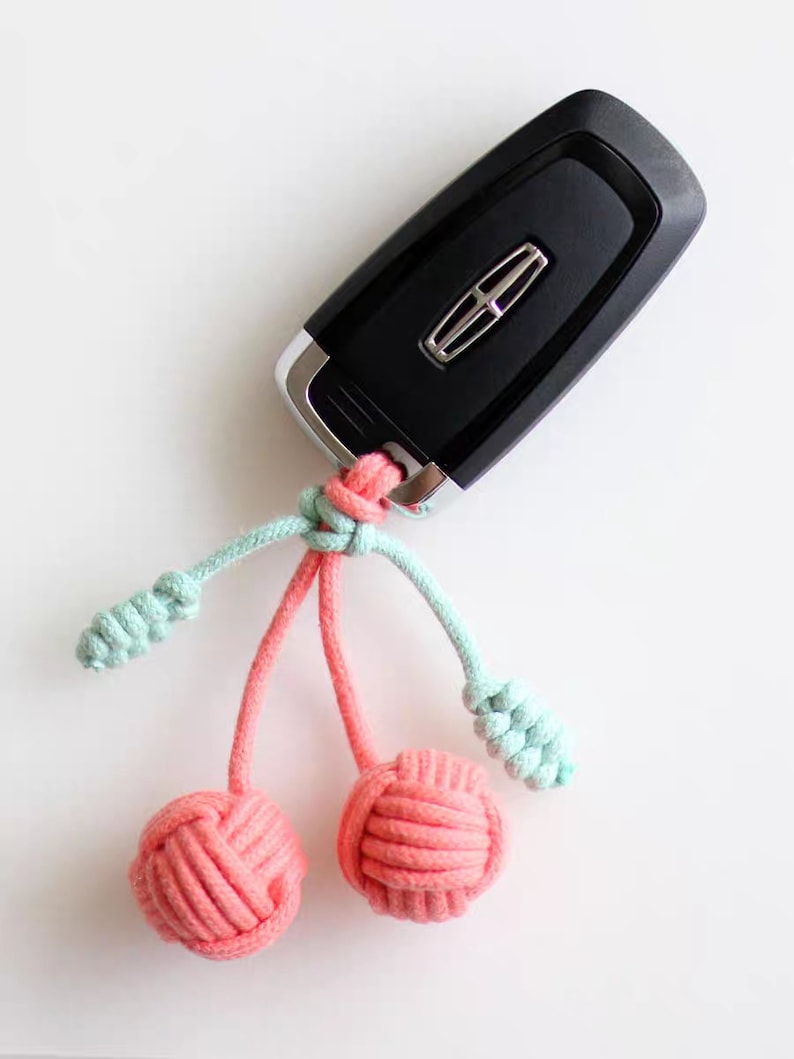 Op de afbeelding: Een zwarte autosleutel met een roze en lichtblauwe touwtjes sleutelhanger met twee roze geknoopte ballen.