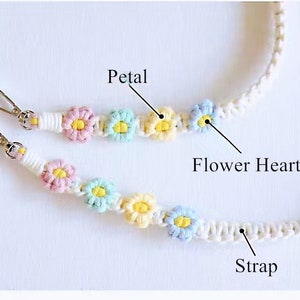 May include: A white crocheted phone strap with colourful flower accents. The strap has two metal clasps and features a white strap with a series of crocheted flowers. Each flower has a yellow centre and pastel pink, blue, and yellow petals. The strap is labelled "Petal", "Flower Heart", and "Strap".