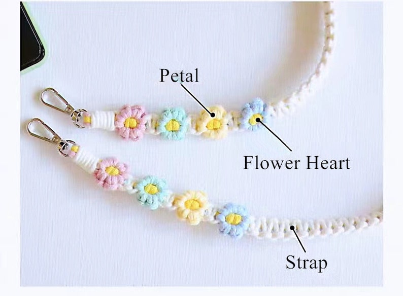 May include: A white crocheted phone strap with colourful flower accents. The strap has two metal clasps and features a white strap with a series of crocheted flowers. Each flower has a yellow centre and pastel pink, blue, and yellow petals. The strap is labelled "Petal", "Flower Heart", and "Strap".