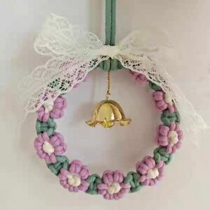 K&ouml;nnte beinhalten: Ein handgefertigter Makramee-Kranz mit rosa und gr&uuml;nen Blumen und einer goldenen Glocke, die in der Mitte h&auml;ngt. Der Kranz ist mit einer wei&szlig;en Schleife aus Spitze verziert.