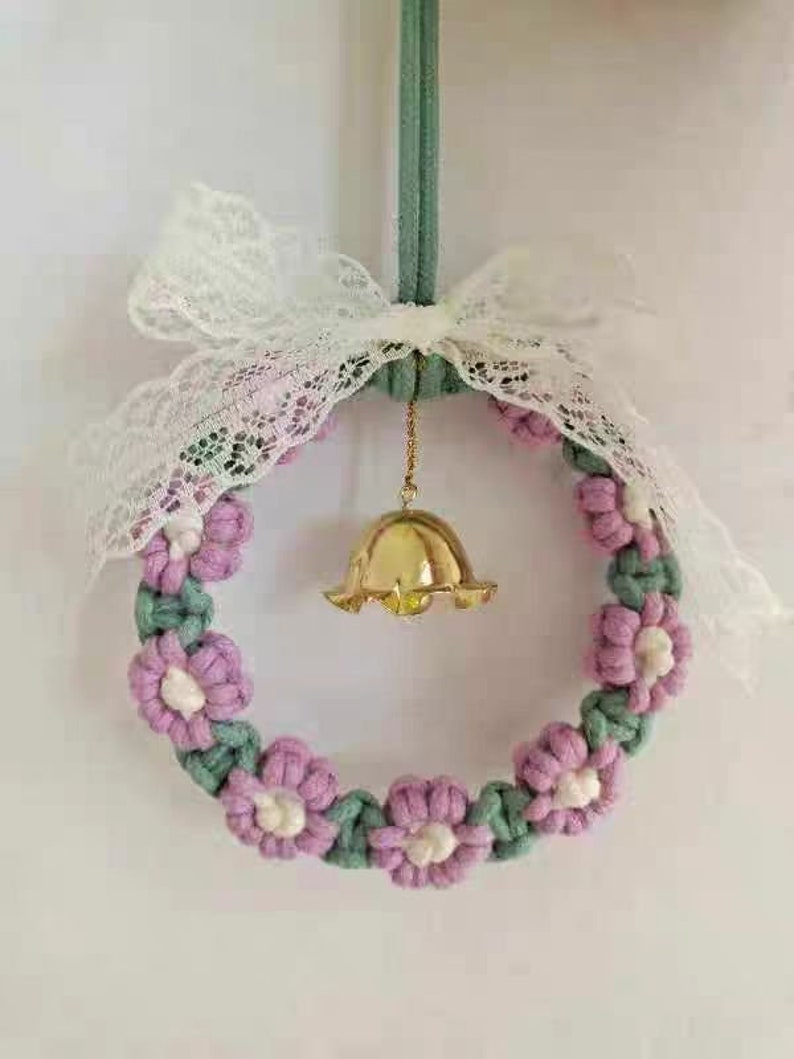 K&ouml;nnte beinhalten: Ein handgefertigter Makramee-Kranz mit rosa und gr&uuml;nen Blumen und einer goldenen Glocke, die in der Mitte h&auml;ngt. Der Kranz ist mit einer wei&szlig;en Schleife aus Spitze verziert.