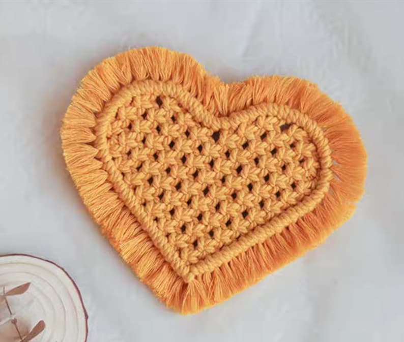 K&ouml;nnte beinhalten: Ein gelber, herzf&ouml;rmiger Makramee-Untersetzer mit Fransenrand. Der Untersetzer ist aus gewebtem Garn gefertigt und hat ein strukturiertes Muster.