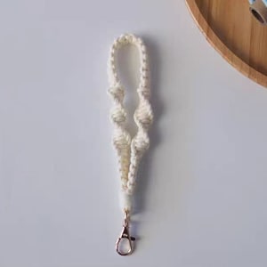 Pode incluir: Um chaveiro de macram&eacute; branco com um fecho de metal dourado. O chaveiro &eacute; feito de uma corda grossa e n&oacute;.