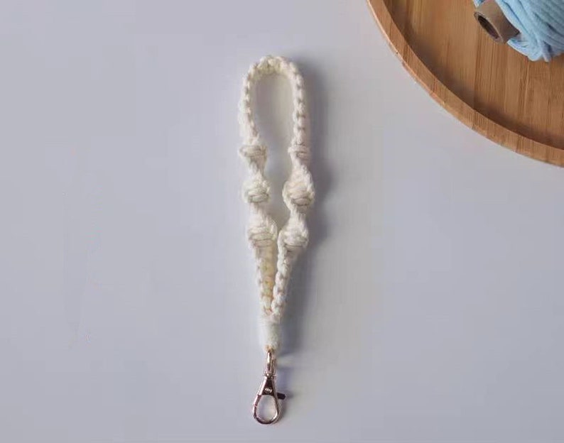 Pode incluir: Um chaveiro de macram&eacute; branco com um fecho de metal dourado. O chaveiro &eacute; feito de uma corda grossa e n&oacute;.