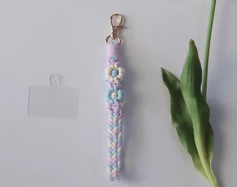 Pu&ograve; includere: Un portachiavi intrecciato rosa, blu e bianco con tre ciondoli a forma di fiore. Il portachiavi ha una chiusura in metallo dorato.