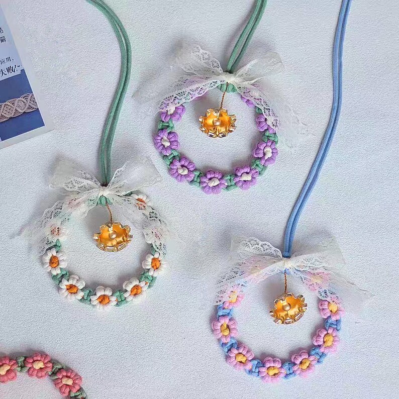 K&ouml;nnte beinhalten: Drei Blumenkr&auml;nze mit goldenen Gl&ouml;ckchen und wei&szlig;en Schleifen aus Spitze auf hellblauem Hintergrund. Die Kr&auml;nze sind aus Garn gefertigt und in den Farben Lila, Gr&uuml;n und Rosa gehalten.