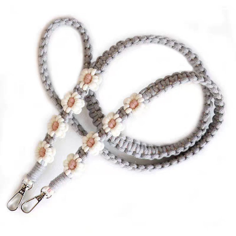May include: A grey braided strap with white and pink daisy-shaped embellishments. The strap has two silver clasps at one end.
