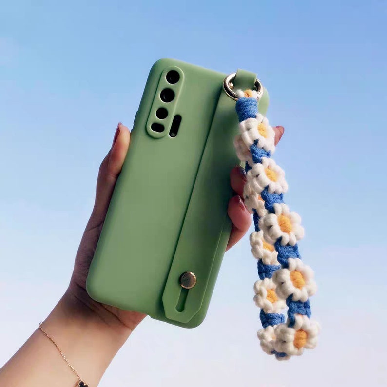 Puede incluir: Una funda de tel&eacute;fono verde con una correa integrada. La correa est&aacute; hecha de hilo blanco y azul y presenta un patr&oacute;n de margarita.