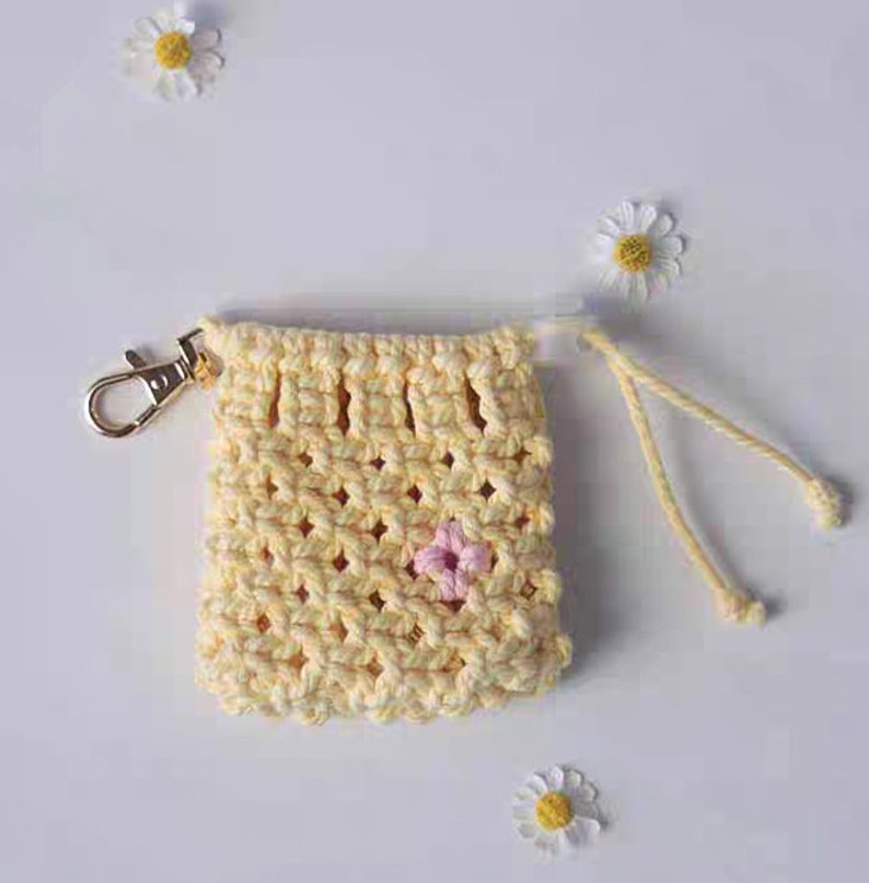 May include: A small yellow crocheted bag with a pink flower design. The bag has a gold metal clasp and a white cord.