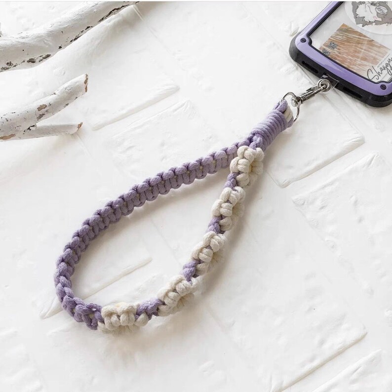 May include: A purple and white braided phone strap with a silver clasp. The strap is made of two different colors of cord, woven together in a pattern.