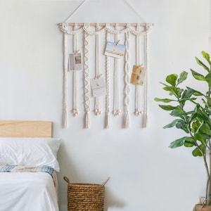 Op de afbeelding: Witte macram&eacute; wandhanger met houten lat en houten kralen. De wandhanger heeft meerdere strengen met verschillende patronen en texturen. Er zijn vijf kleine witte kaartjes met foto's die aan de wandhanger zijn gehangen.