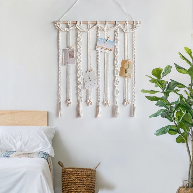 Op de afbeelding: Witte macram&eacute; wandhanger met houten lat en houten kralen. De wandhanger heeft meerdere strengen met verschillende patronen en texturen. Er zijn vijf kleine witte kaartjes met foto's die aan de wandhanger zijn gehangen.
