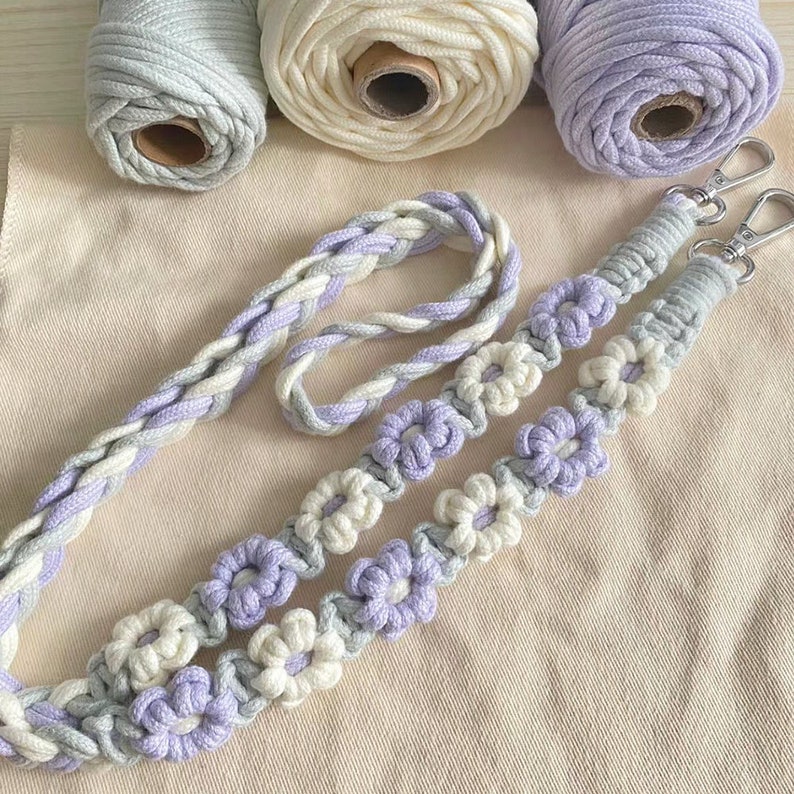 May include: A set of two macrame bag straps with a floral design. The straps are made with a combination of light gray, white, and lavender yarn.
