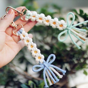 May include: Two handmade macrame keychains. Each keychain features a row of white daisy flowers with yellow centers. The keychains have gold-colored clasps and are accented with blue and white tassels. The background is a soft green.