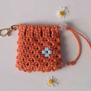 May include: A small orange crocheted drawstring bag with a blue flower design. The bag has a gold metal clasp and a white cord.