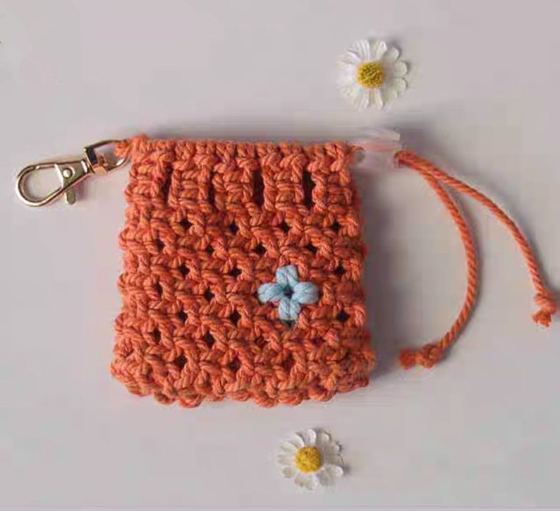 May include: A small orange crocheted drawstring bag with a blue flower design. The bag has a gold metal clasp and a white cord.