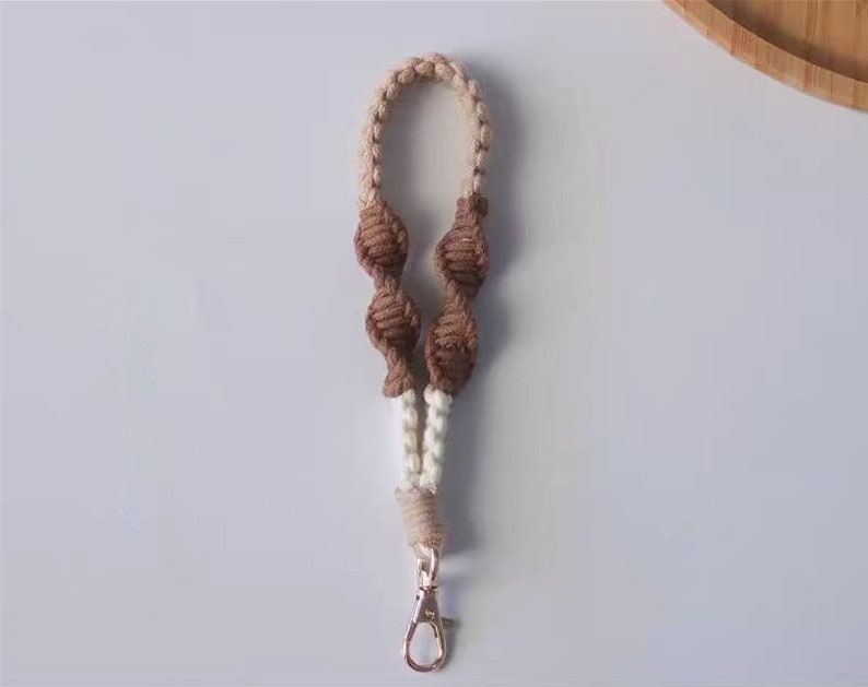 Pode incluir: Um chaveiro tran&ccedil;ado marrom e branco com um fecho de metal. O chaveiro &eacute; feito de dois fios de cores diferentes, com n&oacute;s marrons e fio branco entre os n&oacute;s.