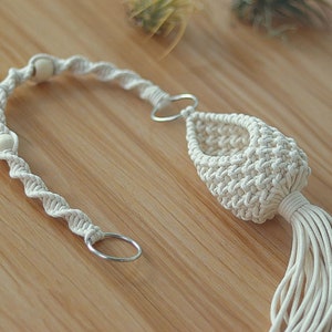 May include: A white macrame plant hanger with wooden beads and a metal ring. The hanger is made of cotton cord and has a tassel at the bottom.