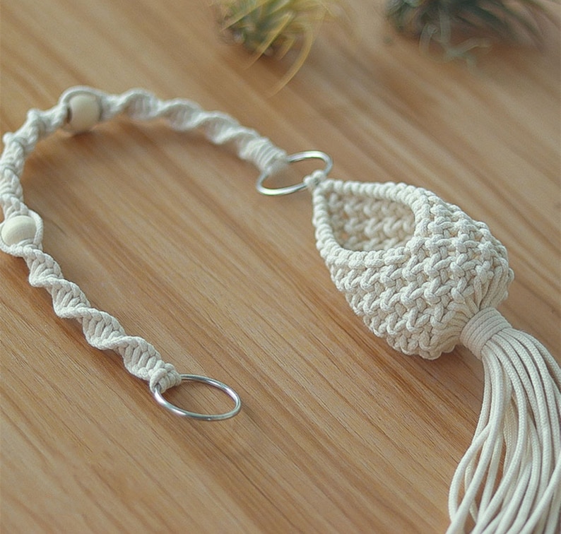 May include: A white macrame plant hanger with wooden beads and a metal ring. The hanger is made of cotton cord and has a tassel at the bottom.