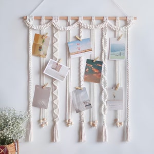May include: White macrame wall hanging with wooden dowel and multiple strands of macrame cord. The wall hanging has a scalloped top and includes eight spaces for displaying photos with wooden pegs.