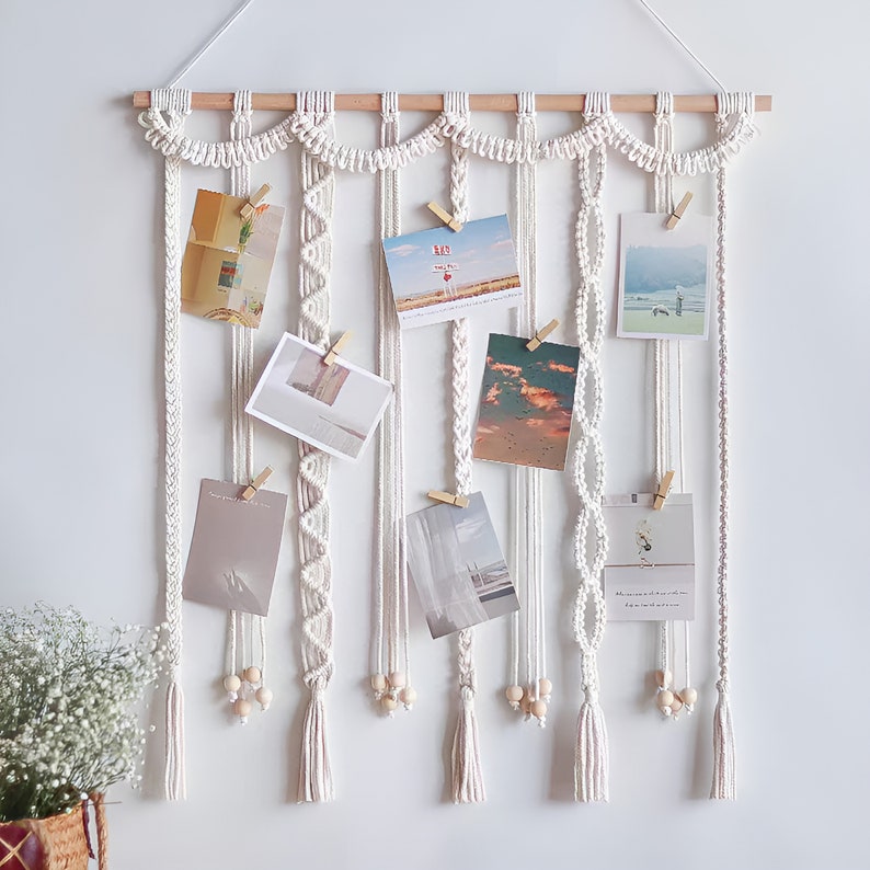 Op de afbeelding: Witte macram&eacute; wandhanger met houten lat en meerdere strengen macram&eacute; koord. De wandhanger heeft een gegolfde bovenkant en bevat acht ruimtes voor het ophangen van foto's met houten wasknijpers.