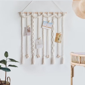 Op de afbeelding: Witte macram&eacute; wandhanger met houten lat en kwastjes. De hanger heeft meerdere strengen macram&eacute; met verschillende patronen en kralen. Er hangen vijf kaarten aan de macram&eacute; met wasknijpers.