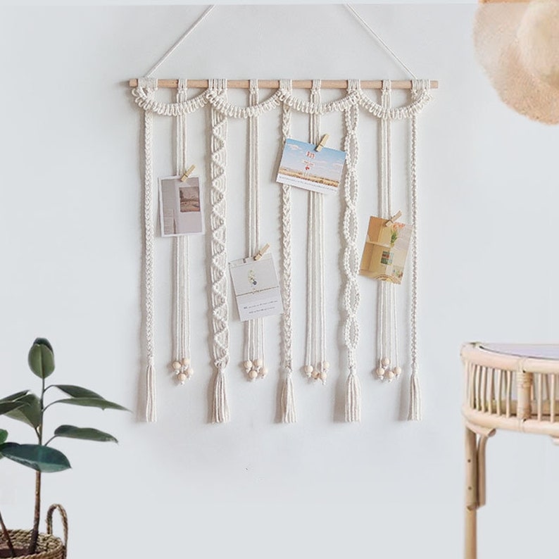Op de afbeelding: Witte macram&eacute; wandhanger met houten lat en kwastjes. De hanger heeft meerdere strengen macram&eacute; met verschillende patronen en kralen. Er hangen vijf kaarten aan de macram&eacute; met wasknijpers.