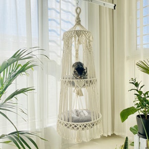 Op de afbeelding: Een witte macramé kattenhangmat met twee niveaus, hangend aan het plafond. Een kat rust op het bovenste niveau. De hangmat bevindt zich voor een raam met witte gordijnen.