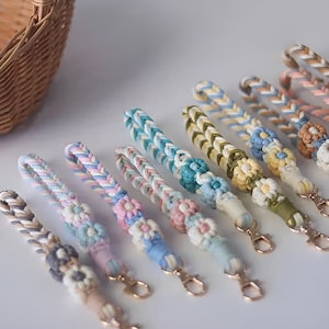 May include: A collection of colourful braided keychains with flower accents. Each keychain features a different colour combination and a small gold clasp.
