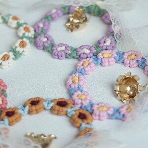 K&ouml;nnte beinhalten: F&uuml;nf geh&auml;kelte Blumenketten mit goldenen Gl&ouml;ckchenanh&auml;ngern. Die Ketten sind in verschiedenen Farben, darunter rosa, blau, gr&uuml;n und wei&szlig;.