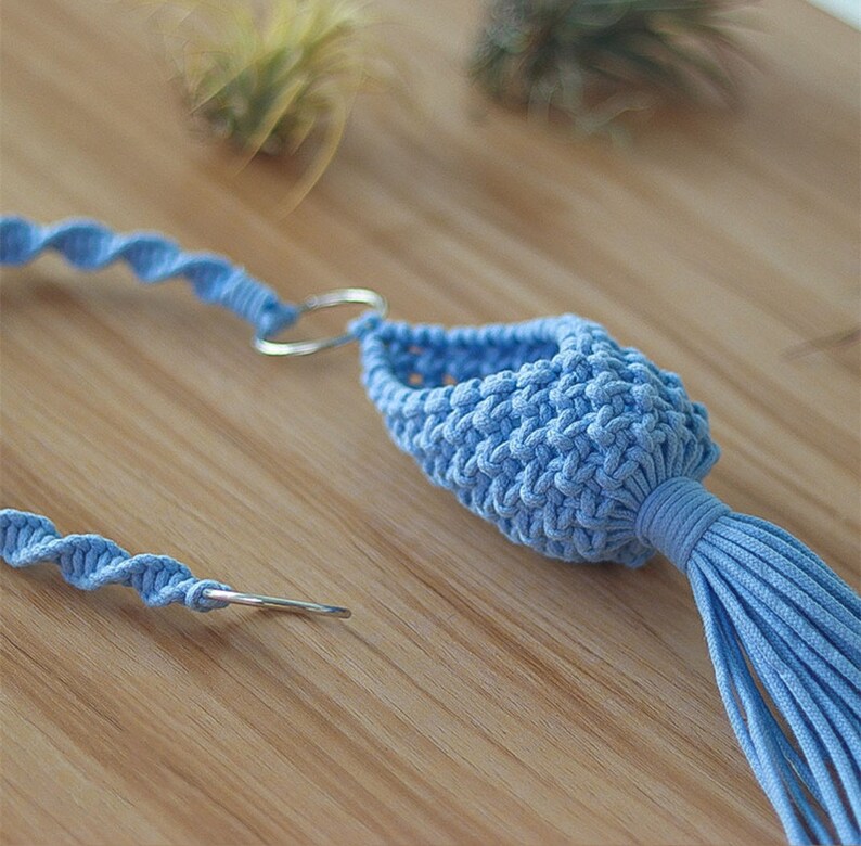 May include: A light blue macrame plant hanger with a metal ring and a tassel. The hanger is made of cotton cord and is designed to hold a small plant.