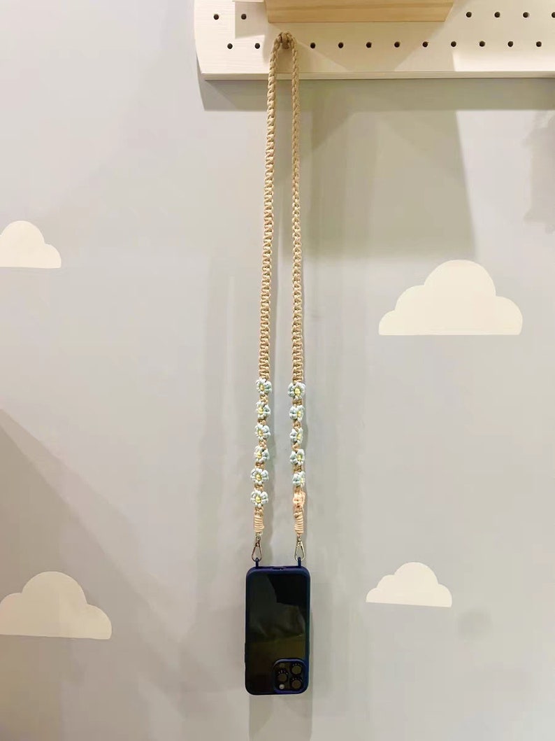 May include: A beige phone strap with blue and white flower accents. The strap is hanging from a pegboard wall.