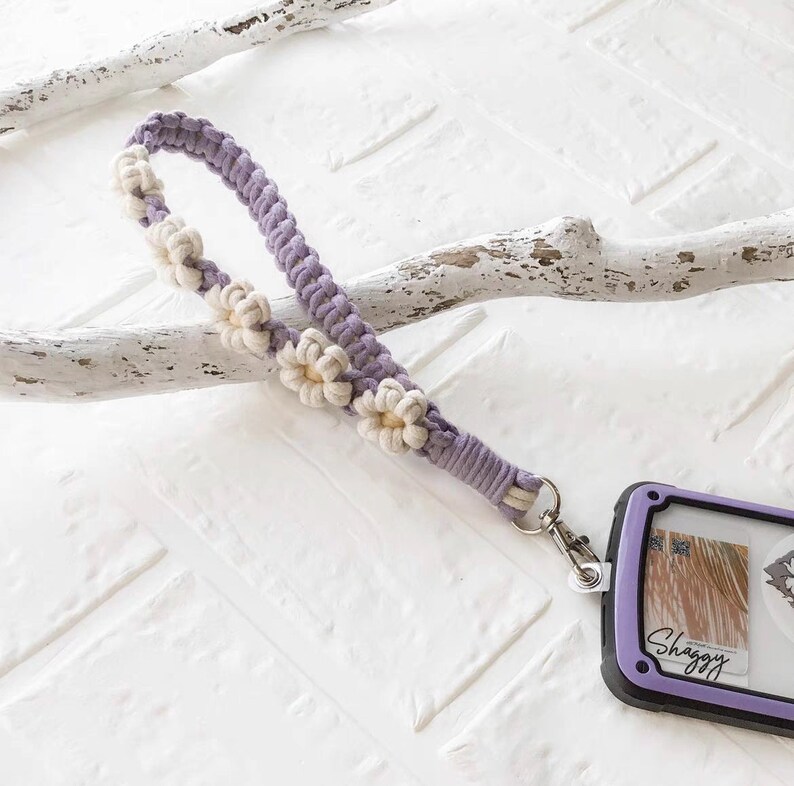 May include: A purple and white crocheted phone strap with a daisy flower design. The strap has a silver metal clip attached to it.