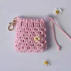 May include: A pink crocheted drawstring bag with a gold metal clasp and a small yellow flower embellishment. The bag has a white background.