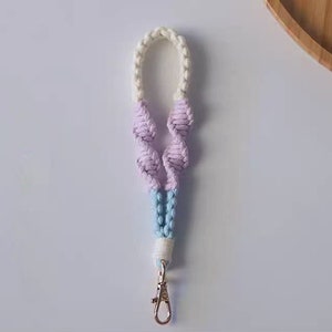 Pode incluir: Um chaveiro de macram&eacute; branco, azul claro e rosa com um fecho prateado.
