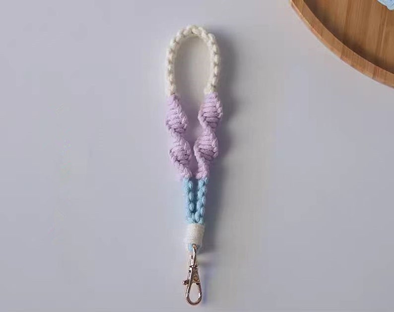 Pode incluir: Um chaveiro de macram&eacute; branco, azul claro e rosa com um fecho prateado.