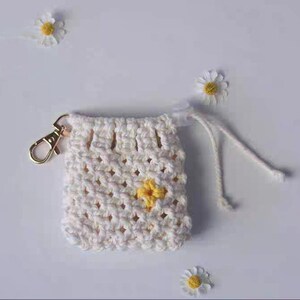 May include: A white crocheted drawstring bag with a yellow flower design and a gold metal clasp. The bag has a white cord.