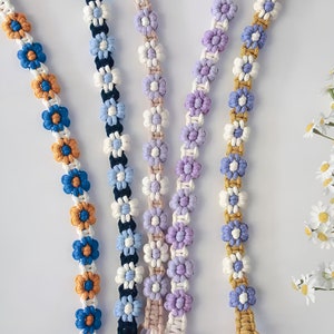 May include: Close-up of several macrame flower chains in various colors, including blue, orange, white, purple, and gold. The daisy-like flowers are evenly spaced along the cords, creating a decorative and handcrafted look. The background is white.