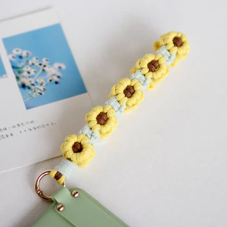 Puede incluir: Una correa para tel&eacute;fono de crochet amarilla y azul claro con un dise&ntilde;o de girasol. La correa tiene un anillo de metal unido a un extremo.