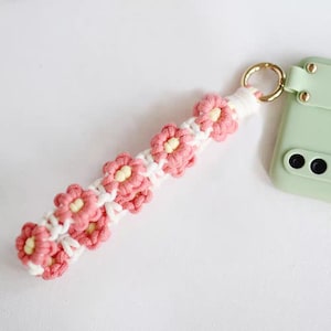 Puede incluir: Una correa de tel&eacute;fono de crochet rosa y blanco con un dise&ntilde;o de flores. La correa est&aacute; unida a una funda de tel&eacute;fono verde.