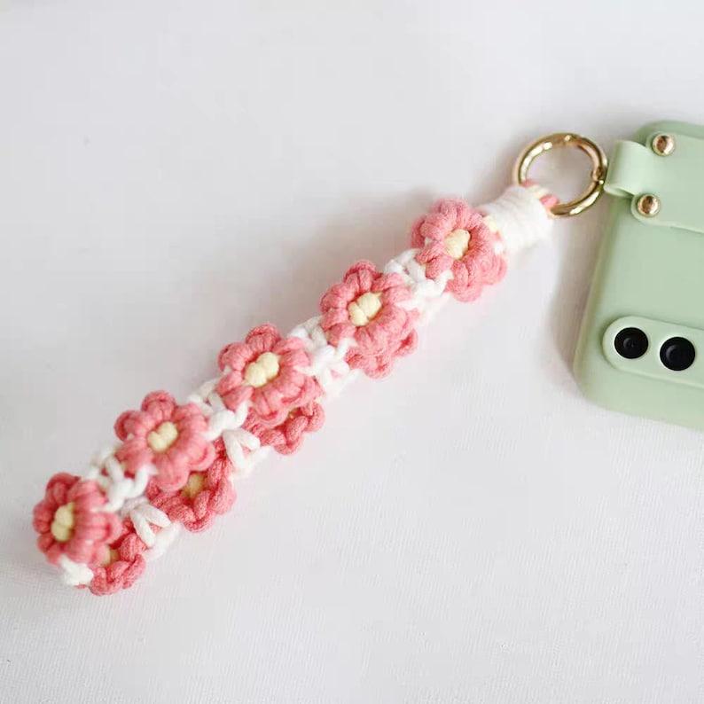 Puede incluir: Una correa de tel&eacute;fono de crochet rosa y blanco con un dise&ntilde;o de flores. La correa est&aacute; unida a una funda de tel&eacute;fono verde.