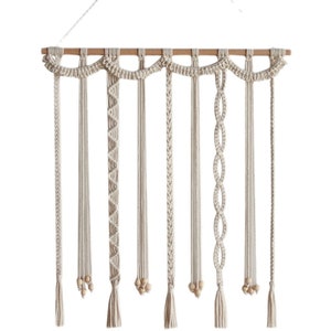 Op de afbeelding: Een macram&eacute; wandhanger met acht strengen van verschillende knopen en texturen. Elke streng heeft een houten kraal en een kwast aan de onderkant. De strengen zijn bevestigd aan een houten lat.