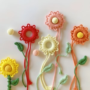 May include: A set of six crocheted flower decorations with colorful yarn. The flowers are in shades of pink, red, yellow, and orange. Each flower has a small bead in the center. The flowers are attached to long strings with green leaves.