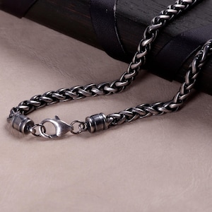 May include: A dark silver-toned wheat chain necklace featuring a polished metallic finish and a sturdy lobster claw clasp. The chain consists of interconnected oval links that create a textured, braided appearance throughout the entire length.