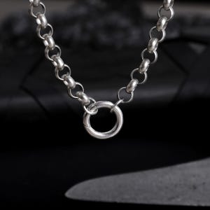 May include: A silver chain necklace with a large circular clasp. The chain is made up of alternating small and large rounded links. The necklace is shown against a dark background, emphasising its metallic shine.