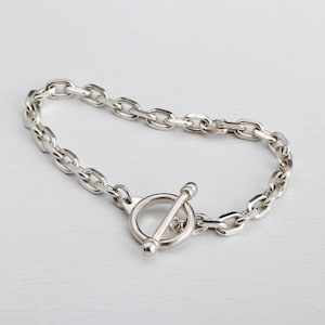 May include: Silver-toned chain bracelet with a toggle clasp. The bracelet features a series of oval links connected together. The toggle clasp is a circular ring with a bar that slides through it to secure the bracelet.
