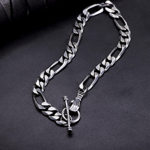 Peut inclure: Un bracelet chaîne argenté avec un fermoir à bascule. Le bracelet présente une série de maillons ovales aplatis et interconnectés. Le fermoir est de type T-bar avec une petite main tenant l'anneau.
